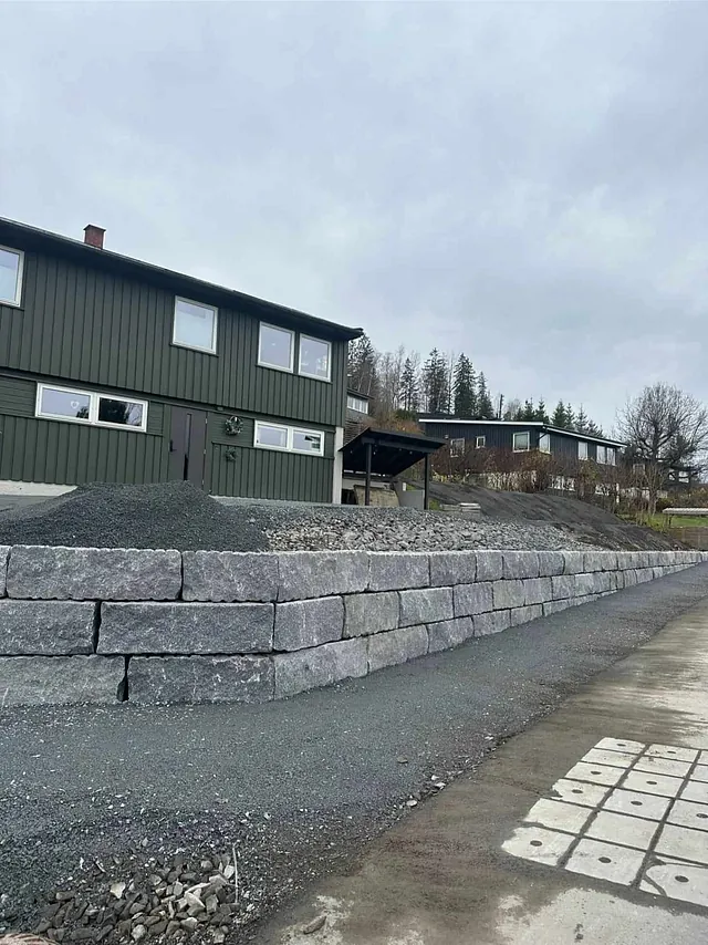 Støttemur av store steinblokker foran grønn enebolig.