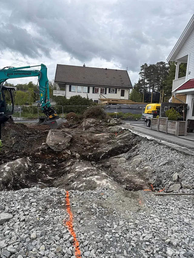 Gravemaskin graver i steinete tomt ved boligområde, omgitt av hus, gjerder og grå himmel.