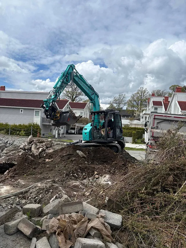 Gravemaskin løfter jord og stein på byggeplass i boligområde med hus og trær rundt.