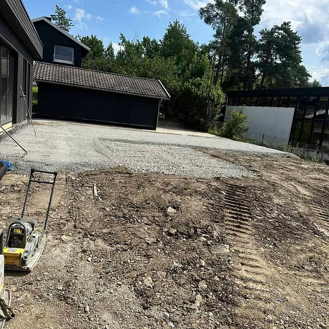 Ferdig gruslagt flate foran mørkt hus og garasje, maskin i forgrunn.